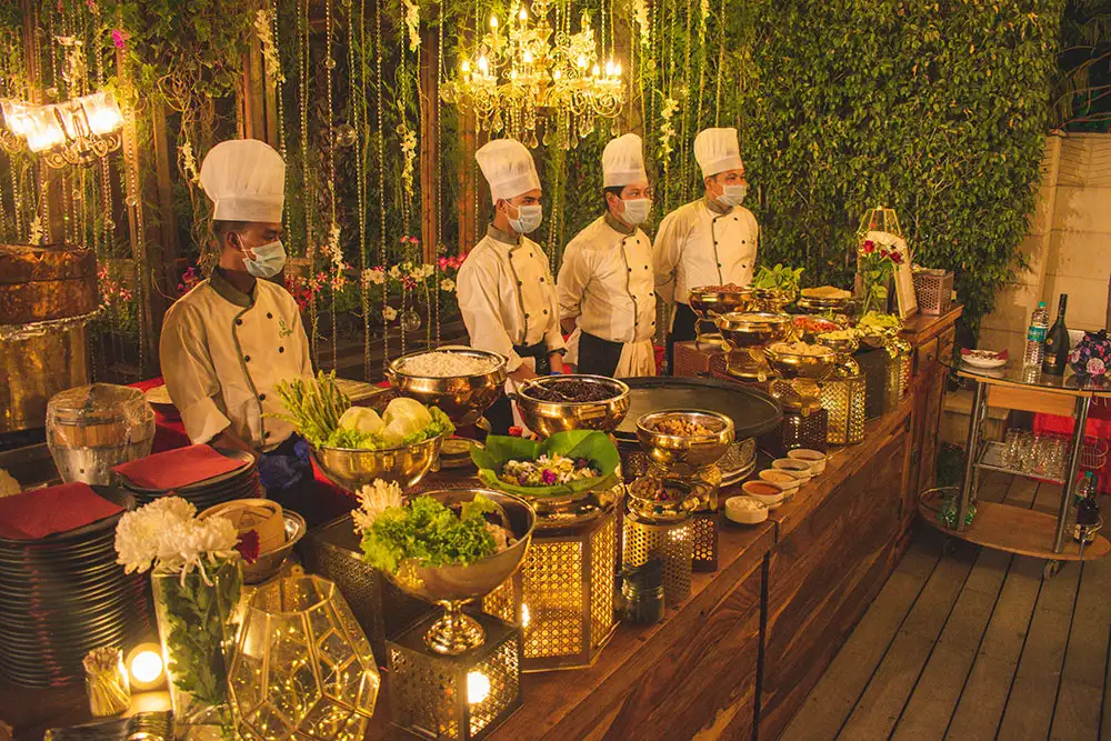Chefs in white uniforms preparing food at a live catering counter in a lit garden venue